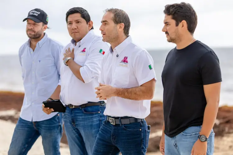 Four men stand together on a beach, with one in a white polo shirt holding a tablet while gesturing toward the ocean and shoreline Four men stand together on a beach, with one in a white polo shirt holding a tablet while gesturing toward the ocean and shoreline
