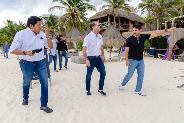 Men stand on a sandy beach near thatched-roof structures while one gestures toward the area, with palm trees and beachfront buildings visible in the background Men stand on a sandy beach near thatched-roof structures while one gestures toward the area, with palm trees and beachfront buildings visible in the background
