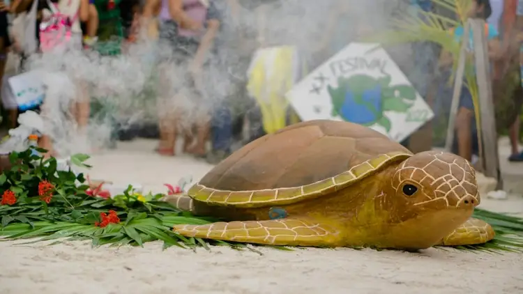 A sea turtle effigy lies on sand surrounded by flowers and greenery, with festival participants visible in the background A sea turtle effigy lies on sand surrounded by flowers and greenery, with festival participants visible in the background