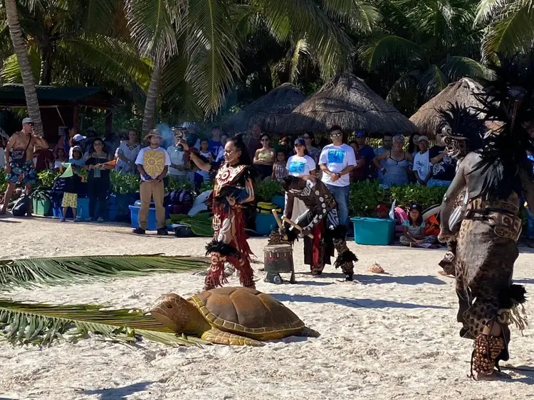 Performers in traditional costumes dance on a beach in front of a sea turtle and spectators gathered under thatched-roof structures Performers in traditional costumes dance on a beach in front of a sea turtle and spectators gathered under thatched-roof structures