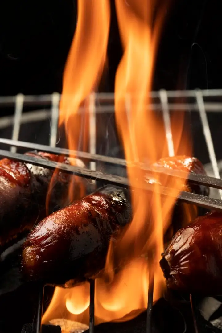 Sausages cook on a metal grate with flames rising above them against a black background Sausages cook on a metal grate with flames rising above them against a black background