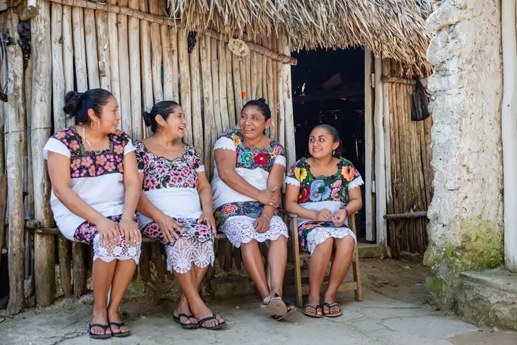 Four women sit together outside a wooden structure with a thatched roof, wearing colorful floral and white patterned clothing
