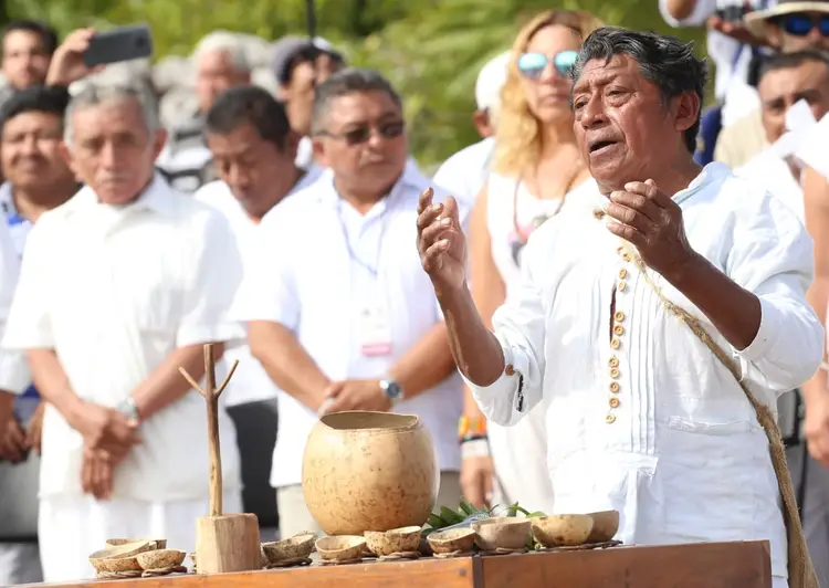 A man in white clothing gestures while speaking to a crowd gathered outdoors, with traditional wooden bowls and vessels displayed on a table in front of him A man in white clothing gestures while speaking to a crowd gathered outdoors, with traditional wooden bowls and vessels displayed on a table in front of him