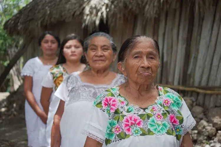 Four women stand outside a thatched-roof structure, with an elderly woman in a embroidered white shirt in the foreground Four women stand outside a thatched-roof structure, with an elderly woman in a embroidered white shirt in the foreground