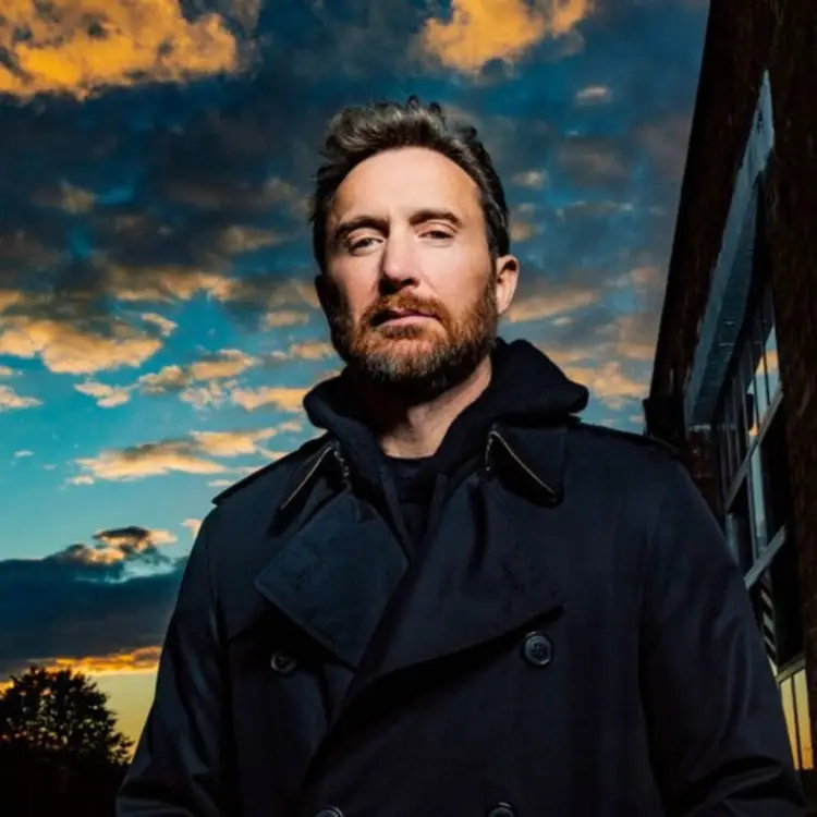 Man with dark hair and beard wearing a black coat stands against a dramatic sunset sky with clouds and a dark structure visible on the right
