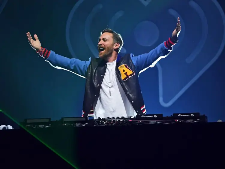 DJ standing behind turntables with arms raised on stage against a blue circular backdrop