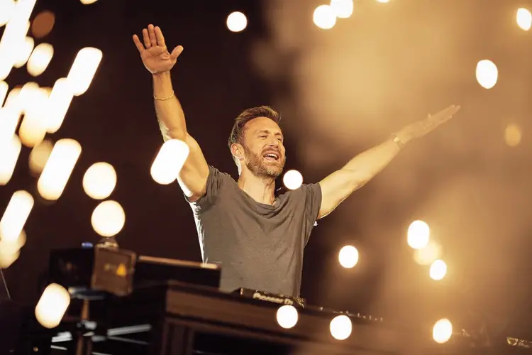Man in a gray shirt raises his arms while performing at a concert with stage lights in the background
