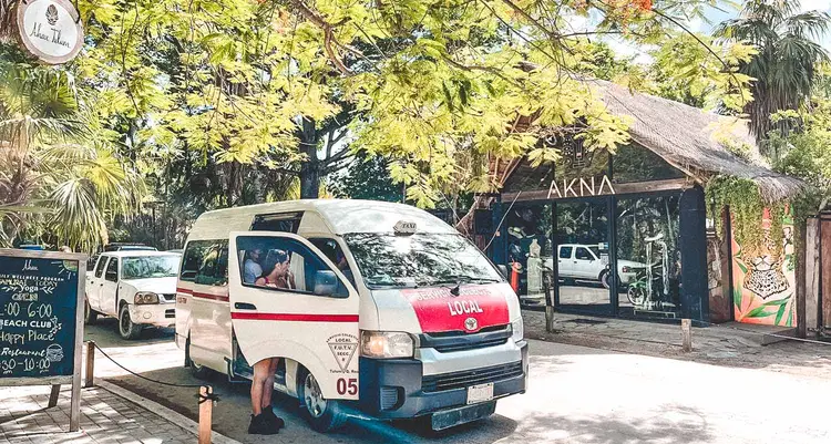 A white and red taxi van with its door open sits parked outside a beachside establishment with thatched-roof structures and palm trees