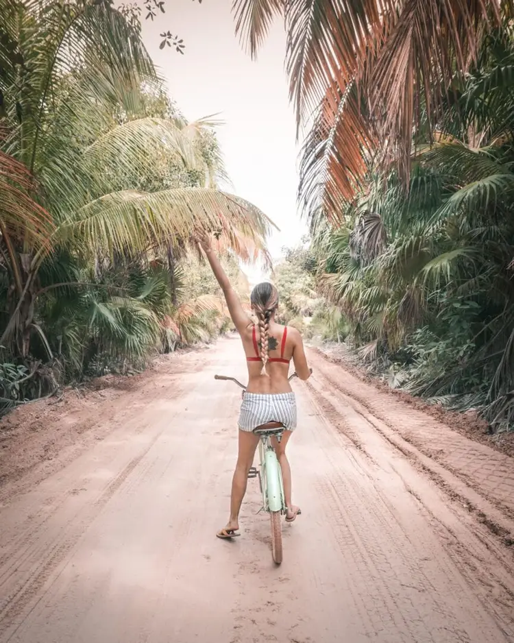 Woman riding a bicycle down a sandy dirt road lined with palm trees, one arm raised