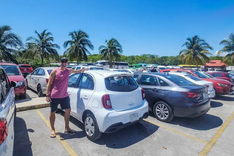 A man in a purple shirt stands between parked cars in a large outdoor lot under clear skies with palm trees in the background