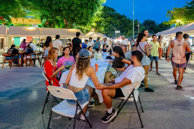Visitors seated at tables and standing among white tents at an outdoor nighttime festival with food vendors and trees lining the street