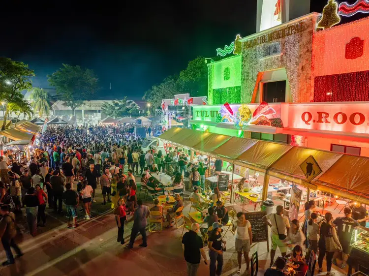 Large crowds gather at a nighttime outdoor festival featuring colorful illuminated vendor stalls and structures in shades of green, orange and red