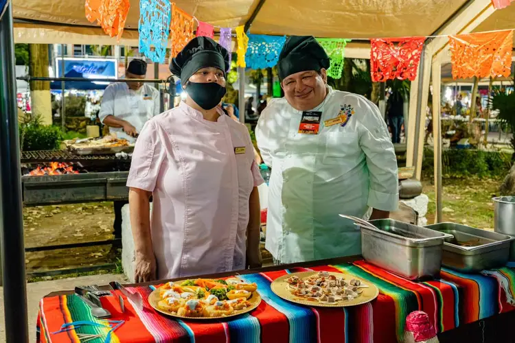 Two cooks stand behind a table with grilled pizzas under a tent decorated with colorful papel picado banners