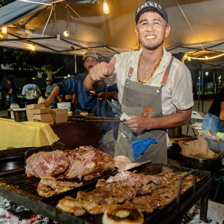 Man in apron and cap grilling meat at an outdoor event under a tent with string lights
