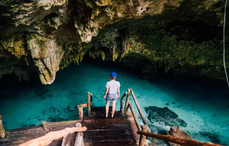 Person standing on wooden dock at edge of turquoise underground cenote pool surrounded by moss-covered limestone cave walls Person standing on wooden dock at edge of turquoise underground cenote pool surrounded by moss-covered limestone cave walls