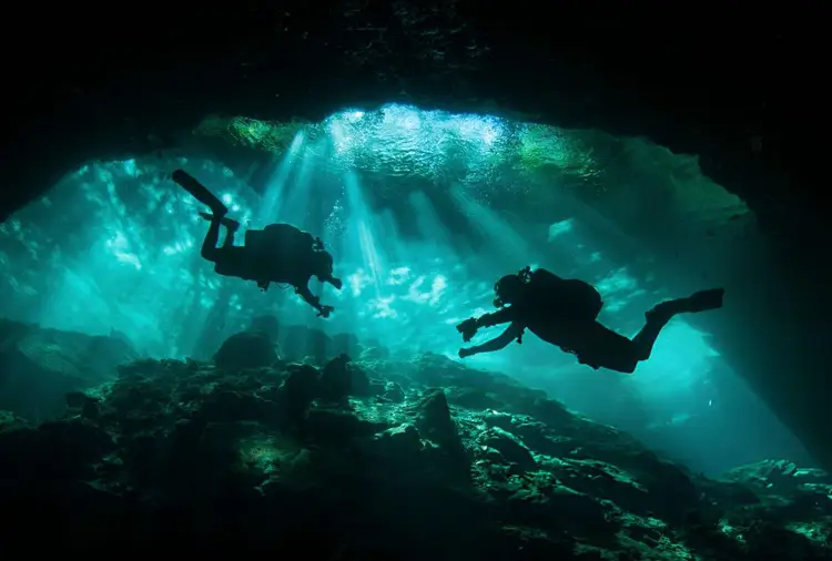 Two scuba divers swim through an underwater cavern with sunlight filtering through the opening above them Two scuba divers swim through an underwater cavern with sunlight filtering through the opening above them