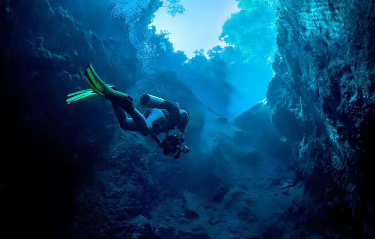 A scuba diver with green fins swims through an underwater cave passage with rocky walls and light filtering from above A scuba diver with green fins swims through an underwater cave passage with rocky walls and light filtering from above