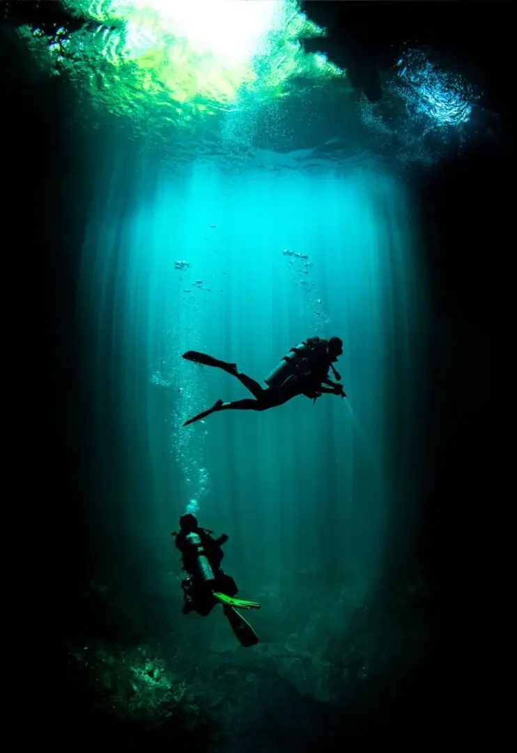 Two scuba divers descend through turquoise water in an underground cenote, with sunlight filtering from the surface above Two scuba divers descend through turquoise water in an underground cenote, with sunlight filtering from the surface above