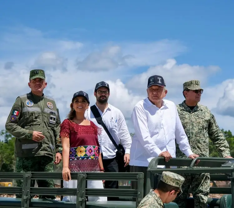 Five people stand on a military vehicle under a blue sky with white clouds, including military personnel in camouflage and uniforms alongside civilians in casua Five people stand on a military vehicle under a blue sky with white clouds, including military personnel in camouflage and uniforms alongside civilians in casua