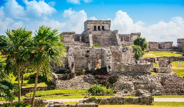 Stone ruins of an ancient pyramid structure surrounded by palm trees and green grounds under a blue sky with white clouds
