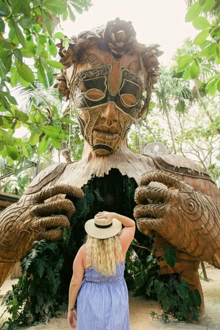 A visitor poses in front of a large wooden sculpture of a skull surrounded by tropical foliage