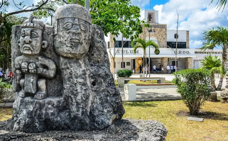 Stone carved pre-Columbian sculptures stand in a plaza with a modern building and palm trees in the background