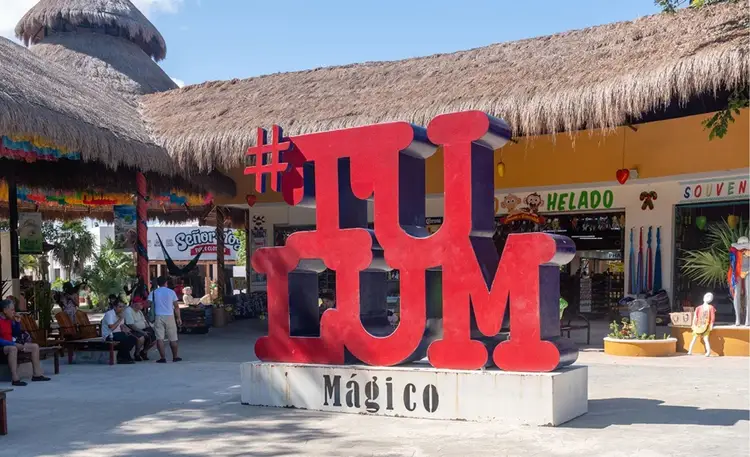 Large red letters spelling a hashtag and Spanish text sit in a plaza surrounded by thatched-roof buildings and souvenir shops