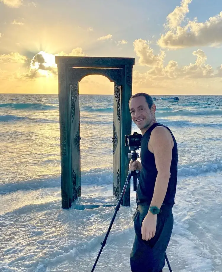 Man standing in ocean waters beside a weathered wooden arch structure, holding a camera on a tripod, with beach and cloudy sky behind him Man standing in ocean waters beside a weathered wooden arch structure, holding a camera on a tripod, with beach and cloudy sky behind him