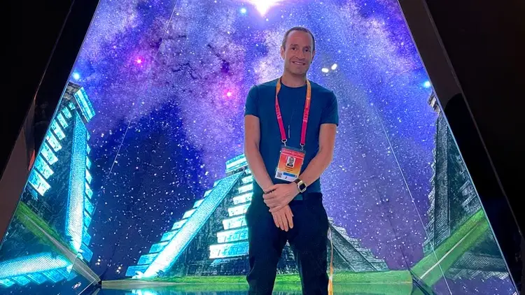 Man wearing a lanyard and dark shirt stands in front of a large illuminated display showing a galaxy and futuristic architecture Man wearing a lanyard and dark shirt stands in front of a large illuminated display showing a galaxy and futuristic architecture