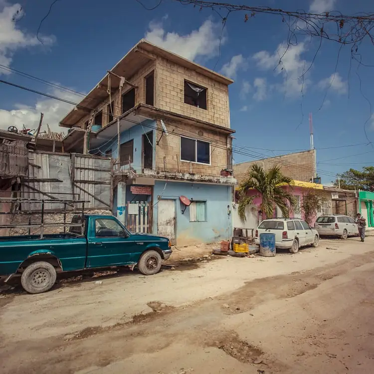 A multi-story residential building with blue lower level and tan upper level stands on a sandy street lined with parked vehicles and neighboring structures A multi-story residential building with blue lower level and tan upper level stands on a sandy street lined with parked vehicles and neighboring structures