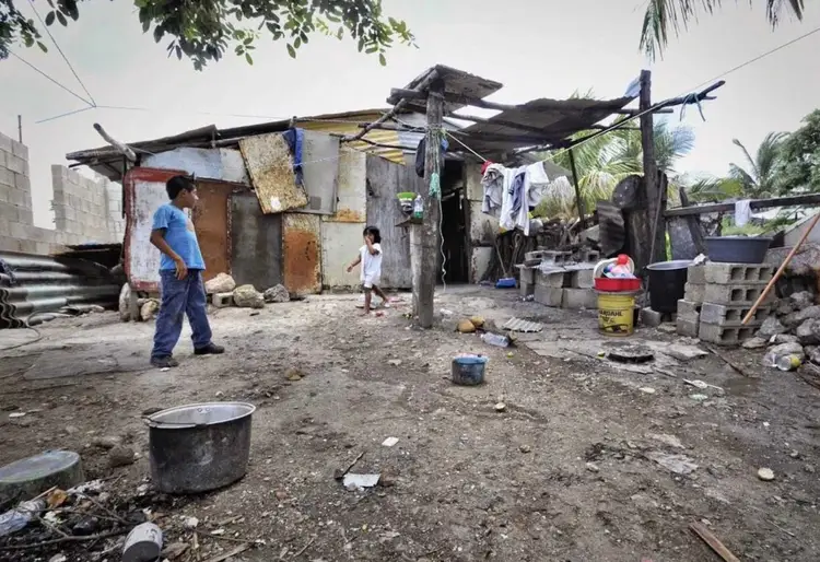 Man in blue shirt stands in a makeshift settlement with basic shelters, cooking vessels, and laundry hanging on lines Man in blue shirt stands in a makeshift settlement with basic shelters, cooking vessels, and laundry hanging on lines