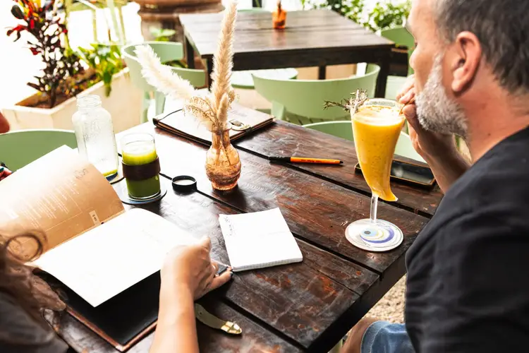 Two people sit at a wooden table with notebooks and beverages during a meeting in a plant-filled outdoor space