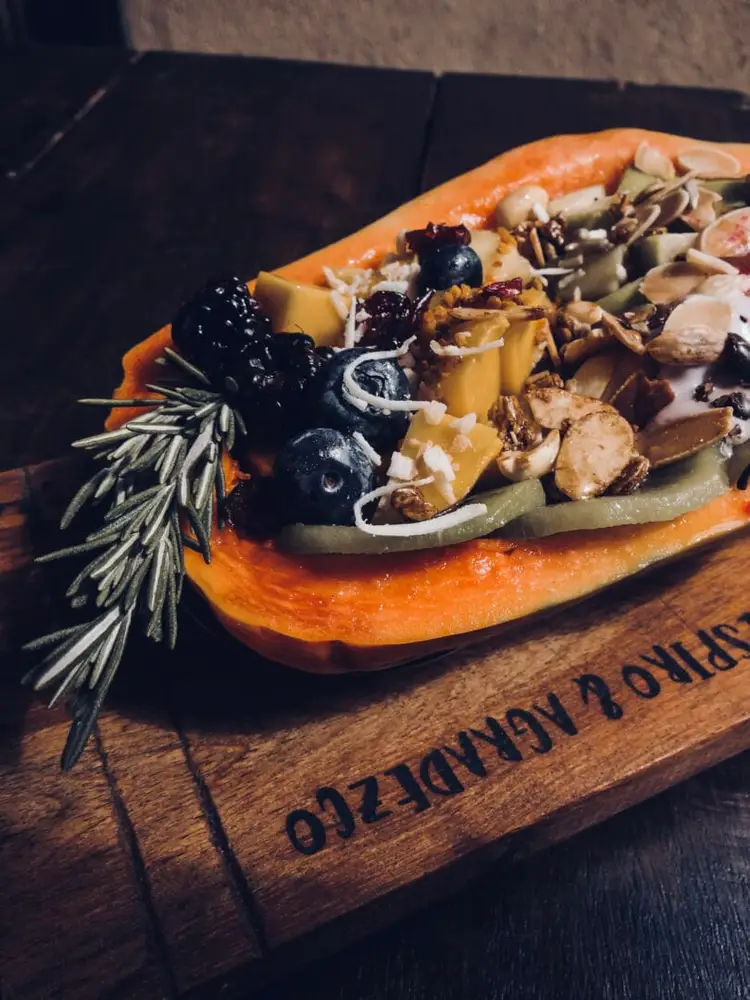 A gourmet sandwich on an orange roll topped with cheese, olives, sliced sausage, onions and rosemary sits on a wooden serving board