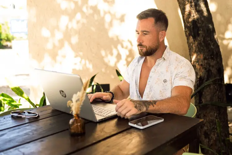 Man working at a laptop at an outdoor table in a shaded courtyard setting