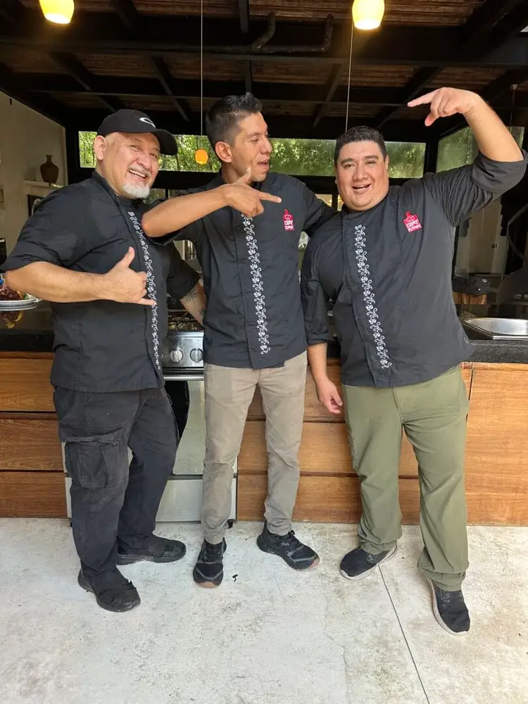 Three men wearing matching black shirts with decorative embroidery laugh and gesture together in a kitchen with wooden countertops and stainless steel appliance Three men wearing matching black shirts with decorative embroidery laugh and gesture together in a kitchen with wooden countertops and stainless steel appliance