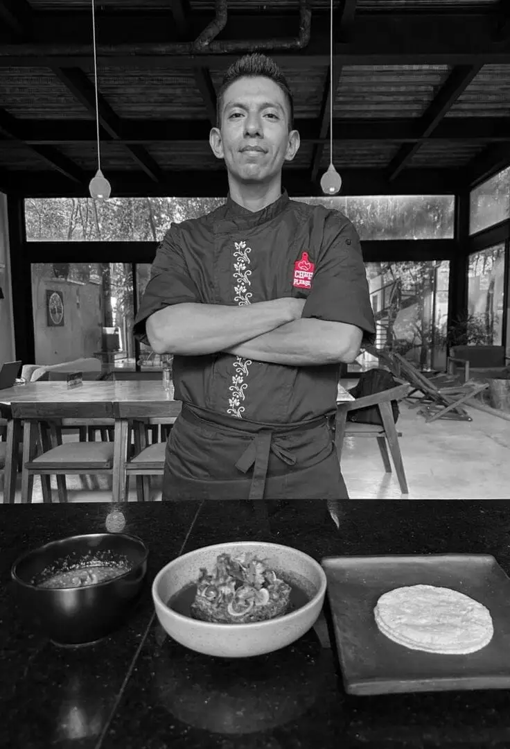 Chef stands with arms crossed behind a counter displaying prepared dishes in bowls and on a plate inside a modern dining space Chef stands with arms crossed behind a counter displaying prepared dishes in bowls and on a plate inside a modern dining space