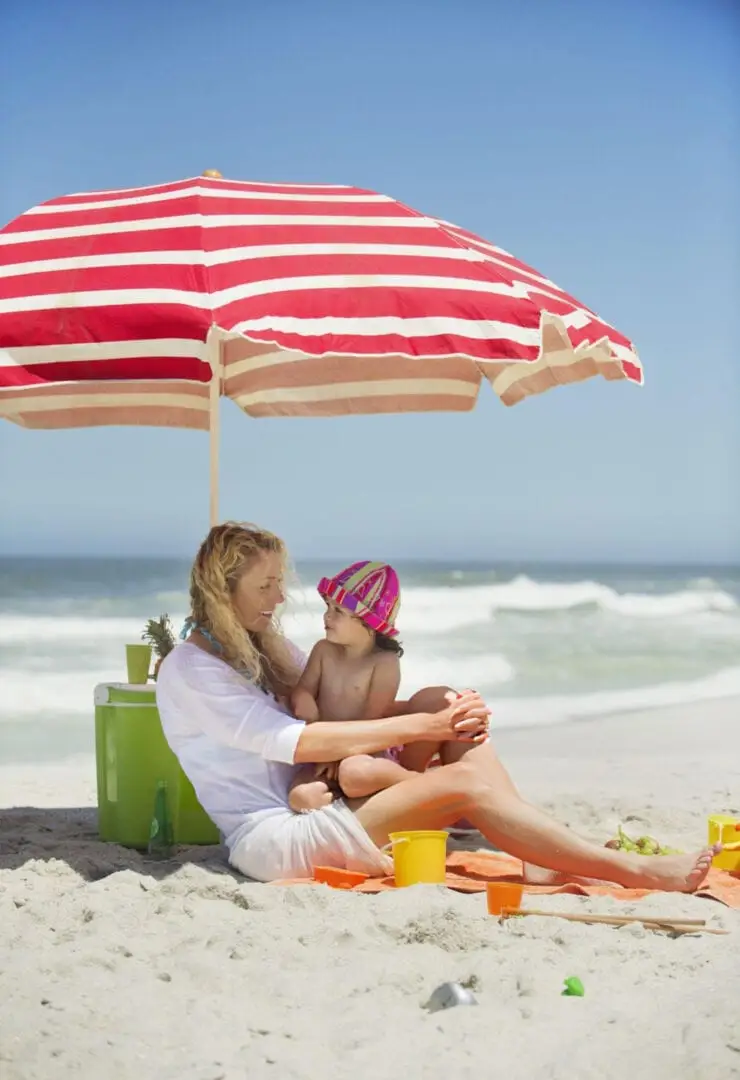 Woman and child sit under a red and white striped umbrella on a sandy beach with ocean waves behind them Woman and child sit under a red and white striped umbrella on a sandy beach with ocean waves behind them