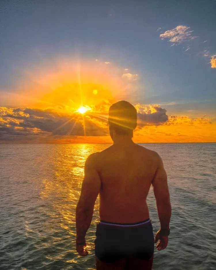 Shirtless man stands in shallow ocean water facing a golden sunset with clouds and sun rays Shirtless man stands in shallow ocean water facing a golden sunset with clouds and sun rays