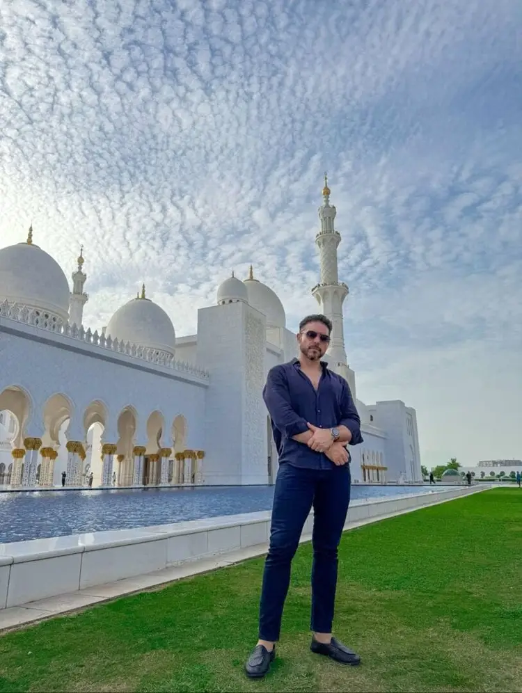 Man in dark blue shirt and sunglasses standing on a lawn in front of a white mosque with domes and minarets Man in dark blue shirt and sunglasses standing on a lawn in front of a white mosque with domes and minarets