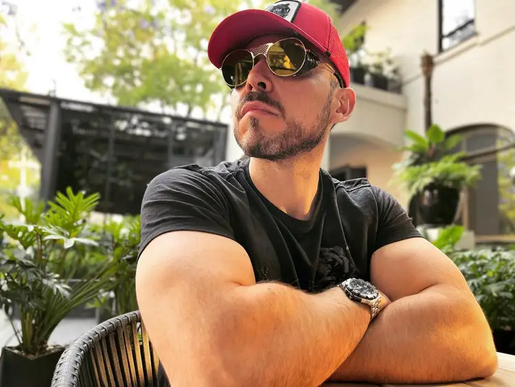 Man wearing a red cap and sunglasses sits at a table with his arms crossed in a courtyard surrounded by potted plants and buildings Man wearing a red cap and sunglasses sits at a table with his arms crossed in a courtyard surrounded by potted plants and buildings