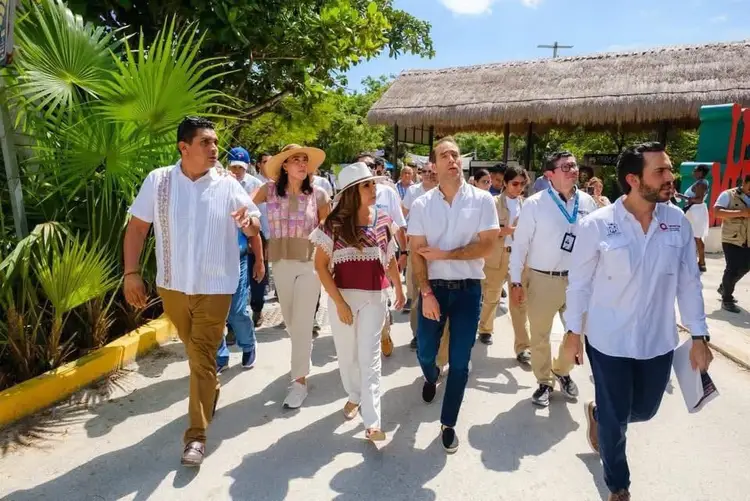 A group of people walk together through a tropical resort area with thatched-roof buildings, palm trees, and a cross visible in the background A group of people walk together through a tropical resort area with thatched-roof buildings, palm trees, and a cross visible in the background