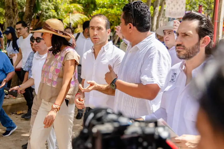 A group of people stand outdoors in a tree-lined area, with several men in light-colored shirts engaged in conversation while others observe and a camera operat A group of people stand outdoors in a tree-lined area, with several men in light-colored shirts engaged in conversation while others observe and a camera operat