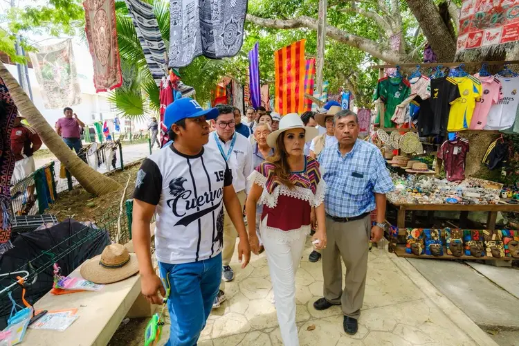Visitors walk through an outdoor market beneath trees with clothing and souvenirs hanging overhead and displayed on tables