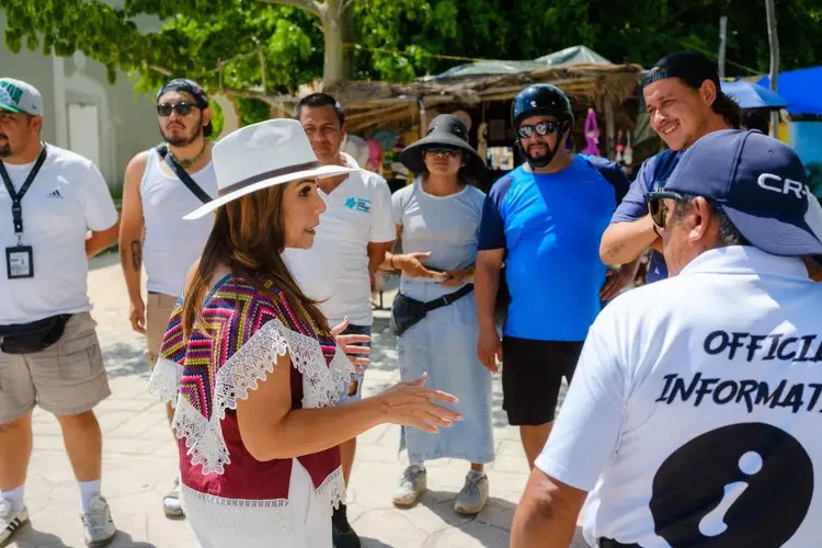 Woman in a white cowboy hat and patterned vest gestures while speaking to a group of people gathered outdoors in a sunny setting