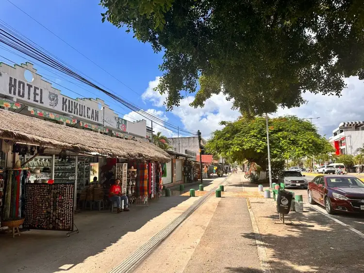 A sandy street lined with small shops and thatched-roof structures, with a hotel sign visible and parked cars along the right side under trees
