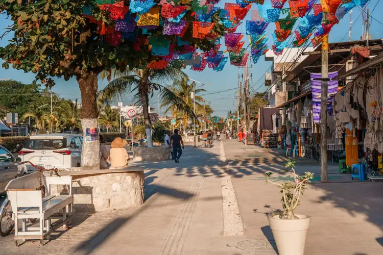 Colorful paper decorations hang above a pedestrian street lined with shops and palm trees, with people walking and sitting in the sunny plaza