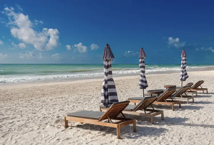 Wooden lounge chairs with gray cushions and striped umbrellas line a sandy beach with turquoise water and white-capped waves under a blue sky