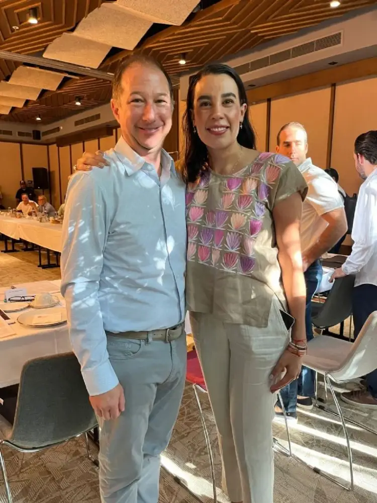 Two people pose together at an indoor event space with wooden ceiling panels and dining tables visible in the background Two people pose together at an indoor event space with wooden ceiling panels and dining tables visible in the background