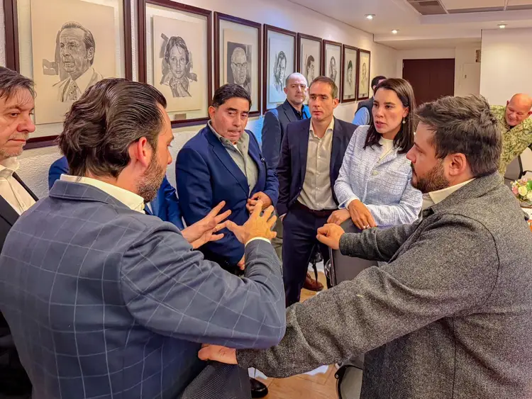 A group of people gather in an indoor meeting space with framed portraits on the wall behind them, appearing to discuss or collaborate on a matter