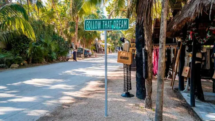 A blue street sign reading "Follow That Dream" stands on a tree-lined pedestrian path with shops and clothing displays on the right side A blue street sign reading "Follow That Dream" stands on a tree-lined pedestrian path with shops and clothing displays on the right side